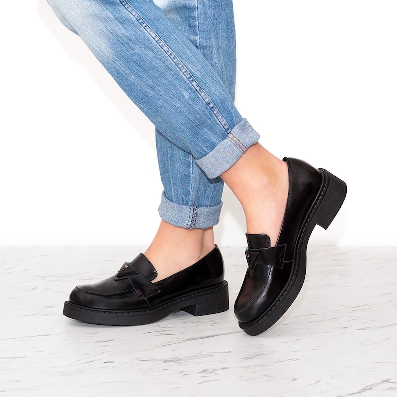 May include: Black leather loafers with a chunky platform sole and a decorative buckle detail. The loafers are worn with rolled up blue jeans.