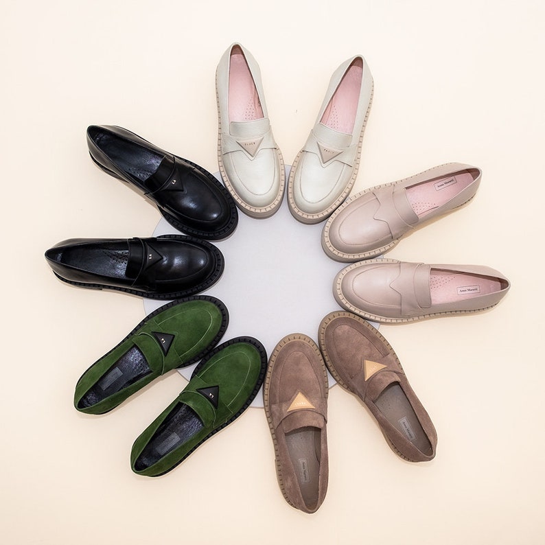 May include: Nine pairs of loafers in various colors and materials, including black leather, green suede, and beige suede. The loafers have a rounded toe and a flat sole.