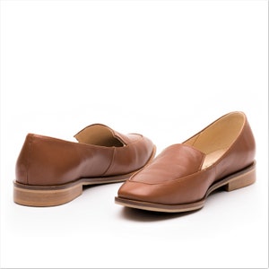 Pu&ograve; includere: Un paio di mocassini in pelle marrone con punta quadrata. Le scarpe hanno un tacco basso e una finitura liscia. I mocassini sono presentati su sfondo bianco, evidenziando il loro stile classico ed elegante.