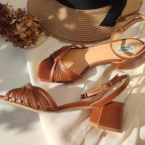 Puede incluir: Sandalias de cuero marr&oacute;n con tac&oacute;n de bloque. Una sandalia presenta un dise&ntilde;o tejido en la puntera, la otra un detalle anudado. Las sandalias se exhiben con un sombrero de paja y flores secas. Altura del tac&oacute;n: aprox. 7 cm.