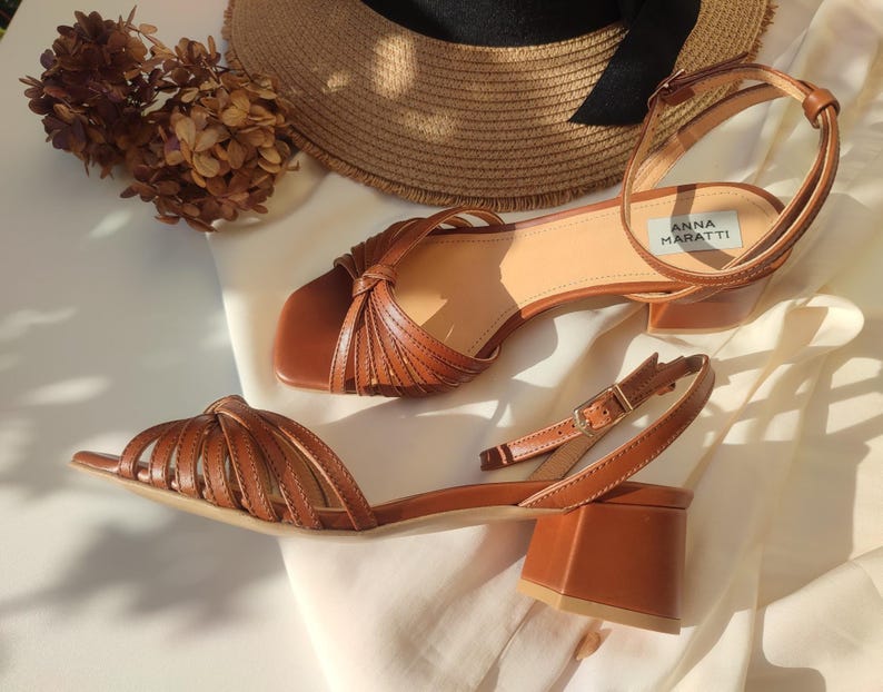 Puede incluir: Sandalias de cuero marr&oacute;n con tac&oacute;n de bloque. Una sandalia presenta un dise&ntilde;o tejido en la puntera, la otra un detalle anudado. Las sandalias se exhiben con un sombrero de paja y flores secas. Altura del tac&oacute;n: aprox. 7 cm.