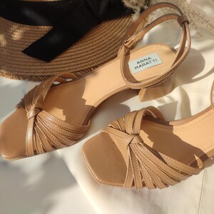 May include: A pair of light brown sandals with a square toe and ankle strap. The sandals have a knotted design on the front and a small block heel. A straw hat with a black ribbon is in the background. The sandals are labeled "ANNA MARATTI".