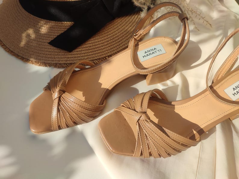 May include: A pair of light brown sandals with a square toe and ankle strap. The sandals have a knotted design on the front and a small block heel. A straw hat with a black ribbon is in the background. The sandals are labeled "ANNA MARATTI".