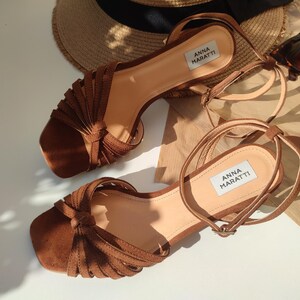 Puede incluir: Sandalias de ante marr&oacute;n con correas en el tobillo. Las sandalias tienen punta cuadrada y llevan el nombre de la marca "ANNA MARATTI". Un sombrero de paja y gafas de sol est&aacute;n en el fondo.
