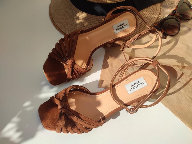 Puede incluir: Sandalias de ante marr&oacute;n con correas en el tobillo. Las sandalias tienen punta cuadrada y llevan el nombre de la marca "ANNA MARATTI". Un sombrero de paja y gafas de sol est&aacute;n en el fondo.