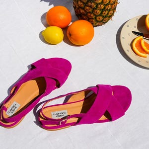 Pode incluir: Sand&aacute;lias f&uacute;csia vibrantes com design de tiras cruzadas, chap&eacute;u de palha, laranjas, um lim&atilde;o e um abacaxi. As sand&aacute;lias t&ecirc;m o texto "ANNA MARATTI" impresso na palmilha.