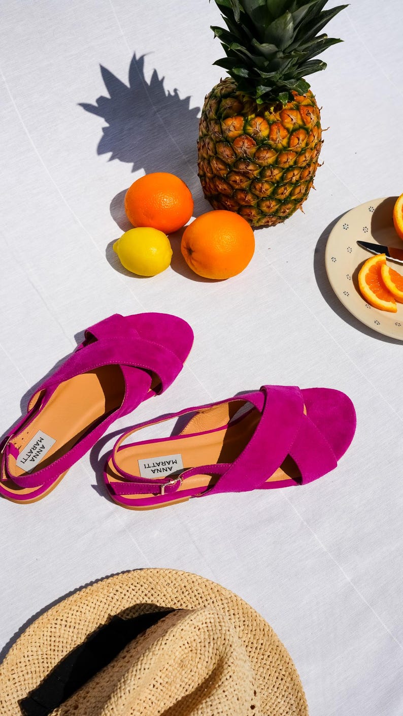 Pode incluir: Sand&aacute;lias f&uacute;csia vibrantes com design de tiras cruzadas, chap&eacute;u de palha, laranjas, um lim&atilde;o e um abacaxi. As sand&aacute;lias t&ecirc;m o texto "ANNA MARATTI" impresso na palmilha.