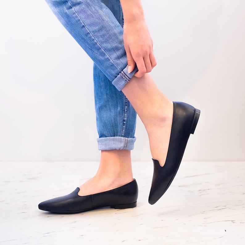 Op de afbeelding: Een paar zwarte leren instapschoenen met spitse neuzen. De schoenen worden gedragen door iemand die een blauwe jeans draagt.