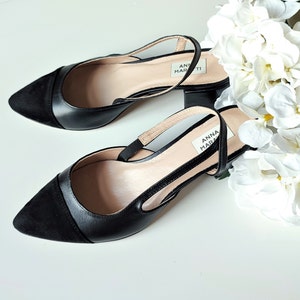 May include: A pair of black slingback heels with a pointed toe and a small block heel. The shoes feature a suede panel on the toe and a leather strap. The brand name "ANNA MARATI" is visible on the insole. White flowers are in the background.