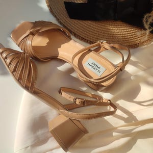 May include: A pair of tan-colored sandals with block heels and ankle straps. The sandals feature a square toe design and multiple straps across the top. The brand name "ANNA MARATTI" is visible on a label inside one of the sandals. A straw hat is in the background.