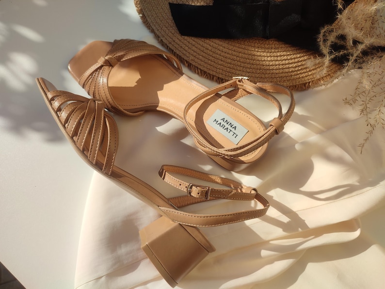 May include: A pair of tan-colored sandals with block heels and ankle straps. The sandals feature a square toe design and multiple straps across the top. The brand name "ANNA MARATTI" is visible on a label inside one of the sandals. A straw hat is in the background.