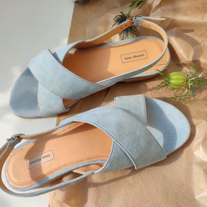 Pode incluir: Um par de sand&aacute;lias de camur&ccedil;a azul claro com design de tiras cruzadas. As sand&aacute;lias t&ecirc;m uma palmilha castanha clara e fecho de fivela. A marca "Anna Maratti" &eacute; vis&iacute;vel na palmilha. Sobre uma superf&iacute;cie castanha.