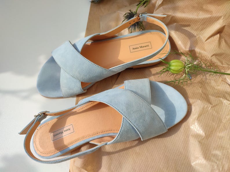 Pode incluir: Um par de sand&aacute;lias de camur&ccedil;a azul claro com design de tiras cruzadas. As sand&aacute;lias t&ecirc;m uma palmilha castanha clara e fecho de fivela. A marca "Anna Maratti" &eacute; vis&iacute;vel na palmilha. Sobre uma superf&iacute;cie castanha.