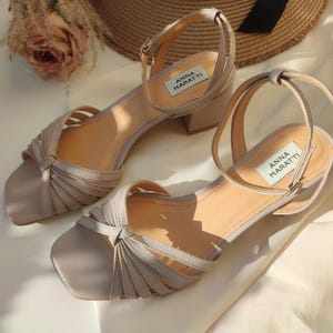May include: A pair of light gray sandals with a square toe and a small block heel. The sandals have multiple straps across the top and an ankle strap. The brand name "ANNA MARATTI" is visible on a label on each shoe.