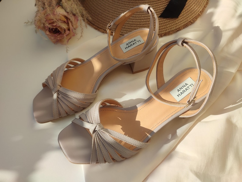 May include: A pair of light gray sandals with a square toe and a small block heel. The sandals have multiple straps across the top and an ankle strap. The brand name "ANNA MARATTI" is visible on a label on each shoe.