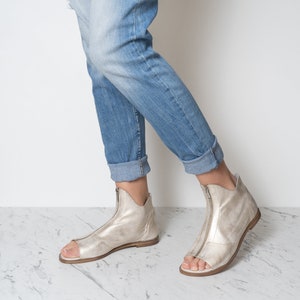 Op de afbeelding: Een paar zilveren leren sandalen met een rits aan de voorkant. De sandalen worden gedragen door iemand die een blauwe jeans draagt.