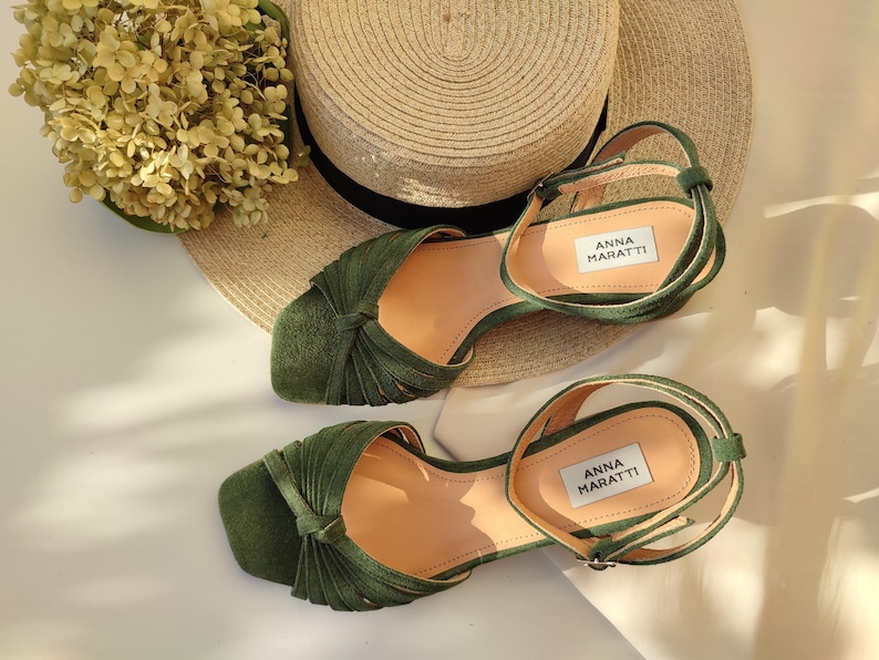 Może przedstawiać: Para oliwkowych sandał&oacute;w z wiązanym wzorem na palcach i paskami na kostkach. Obok znajduje się słomkowy kapelusz i suszone kwiaty. Na wkładce widnieje nazwa marki "ANNA MARATTI".