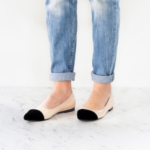 May include: A pair of beige ballet flats with black toe caps. The flats are worn by a person wearing blue jeans.