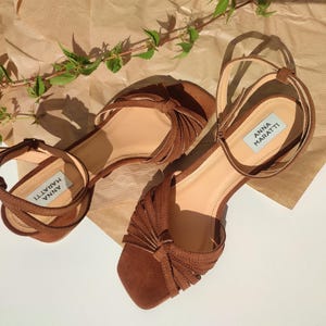 Puede incluir: Un par de sandalias de ante marr&oacute;n con correas en el tobillo y un dise&ntilde;o anudado en las correas de los dedos. Las sandalias tienen la marca "ANNA MARATTI" en la plantilla. Se encuentran sobre una superficie marr&oacute;n claro con follaje verde.