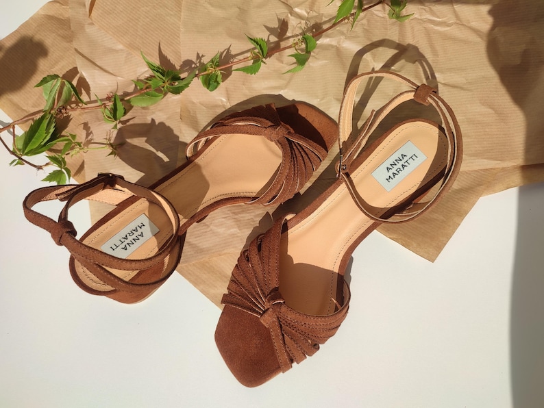 Puede incluir: Un par de sandalias de ante marr&oacute;n con correas en el tobillo y un dise&ntilde;o anudado en las correas de los dedos. Las sandalias tienen la marca "ANNA MARATTI" en la plantilla. Se encuentran sobre una superficie marr&oacute;n claro con follaje verde.