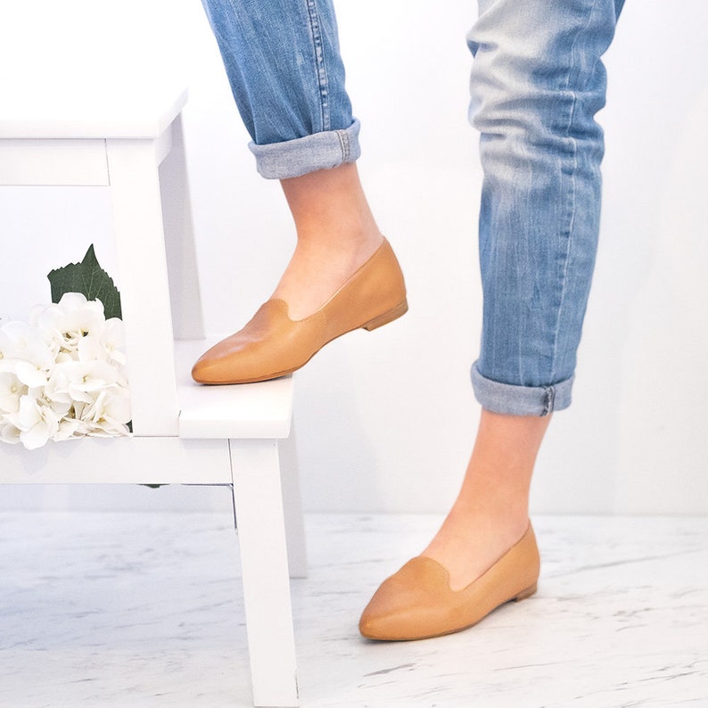 Op de afbeelding: Een paar beige leren spitse ballerina's met een lage hak. De schoenen worden gedragen door iemand die een blauwe jeans draagt.