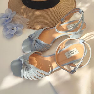 Może przedstawiać: Jasnoniebieskie sandały z paskami na kostkę i wiązanym wzorem. Sandały mają niski obcas, a na wkładce widnieje nazwa marki "ANNA MARATTI". Na zdjęciu widoczny jest r&oacute;wnież słomkowy kapelusz i niebieskie kwiatowe akcesoria do włos&oacute;w.