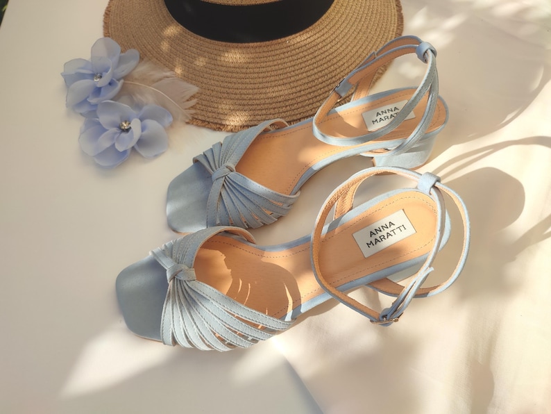Może przedstawiać: Jasnoniebieskie sandały z paskami na kostkę i wiązanym wzorem. Sandały mają niski obcas, a na wkładce widnieje nazwa marki "ANNA MARATTI". Na zdjęciu widoczny jest r&oacute;wnież słomkowy kapelusz i niebieskie kwiatowe akcesoria do włos&oacute;w.