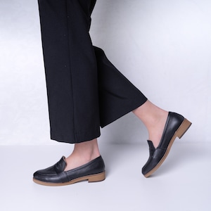 Könnte beinhalten: Ein Paar schwarze Leder-Slipper mit brauner Sohle. Die Slipper haben ein schlichtes Design und werden von einer Person in schwarzen Hosen getragen.