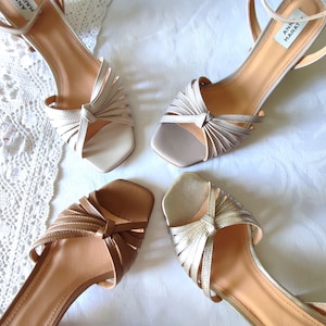 May include: Four pairs of women's sandals in various neutral tones. The sandals feature thin straps, open toes, and decorative knotted details. The soles are a light tan color, and the heels are low. The sandals are arranged on a white surface.