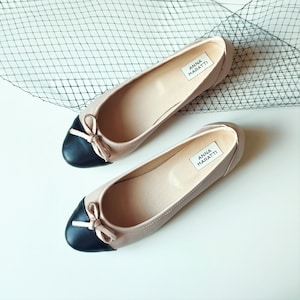 May include: A pair of beige ballet flats with black toe caps and bows. The shoes have the brand name "ANNA MARATTI" printed on the insole.
