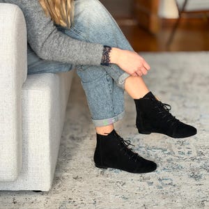 Könnte beinhalten: Schwarze Stiefeletten aus Wildleder mit Schnürverschluss. Die Stiefel haben einen niedrigen Absatz und werden mit einer blauen Jeans getragen. Die Stiefel stehen auf einem gemusterten Teppich. Die Person sitzt auf einem hellen Sofa.