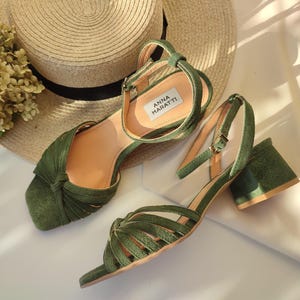 Może przedstawiać: Sandały zamszowe w kolorze oliwkowej zieleni z obcasem w kształcie klocka. Sandały mają pasek wok&oacute;ł kostki i wiązany wz&oacute;r z przodu. Marka "ANNA MARATTI" jest widoczna na wkładce. Słomkowy kapelusz i suszone kwiaty są w tle.