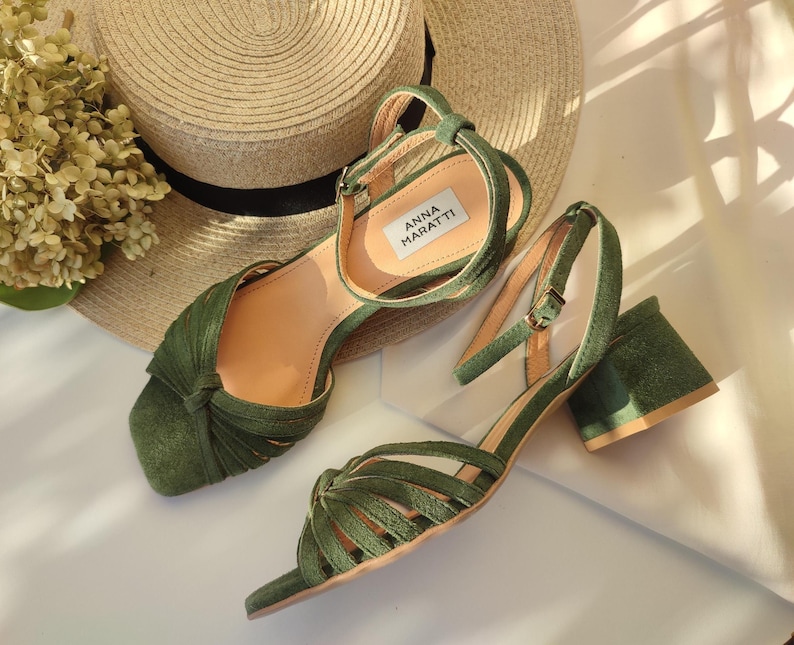 Może przedstawiać: Sandały zamszowe w kolorze oliwkowej zieleni z obcasem w kształcie klocka. Sandały mają pasek wok&oacute;ł kostki i wiązany wz&oacute;r z przodu. Marka "ANNA MARATTI" jest widoczna na wkładce. Słomkowy kapelusz i suszone kwiaty są w tle.