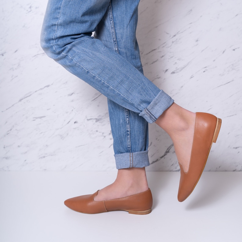 Op de afbeelding: Een paar bruine leren ballerina's met spitse neuzen. De schoenen worden gedragen door iemand die een blauwe jeans draagt.