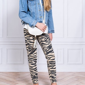 May include: A young person wearing a light blue denim jacket, a white t-shirt, and brown and white zebra print leggings. They are carrying a white handbag with a tassel and wearing light brown lace-up shoes.