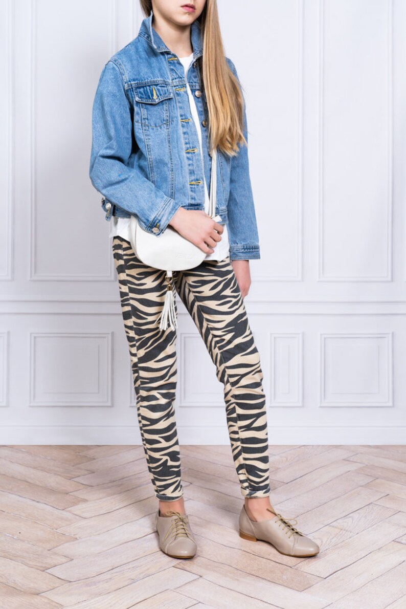 May include: A young person wearing a light blue denim jacket, a white t-shirt, and brown and white zebra print leggings. They are carrying a white handbag with a tassel and wearing light brown lace-up shoes.
