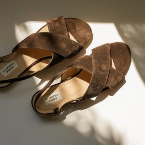 May include: Two brown suede sandals with a crisscross strap design. The sandals feature a low heel and a buckle fastening. The brand name "ANNA MARATTI" is printed on the insole. They are placed on a white surface with shadows.