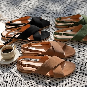 Pode incluir: Uma variedade de sand&aacute;lias de camur&ccedil;a em preto, castanho e verde, com tiras cruzadas e tiras no tornozelo. As sand&aacute;lias est&atilde;o dispostas sobre uma manta estampada, juntamente com uma ch&aacute;vena de caf&eacute; e um livro aberto.