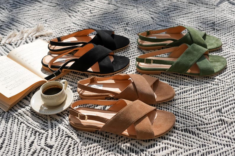 Pode incluir: Uma variedade de sand&aacute;lias de camur&ccedil;a em preto, castanho e verde, com tiras cruzadas e tiras no tornozelo. As sand&aacute;lias est&atilde;o dispostas sobre uma manta estampada, juntamente com uma ch&aacute;vena de caf&eacute; e um livro aberto.