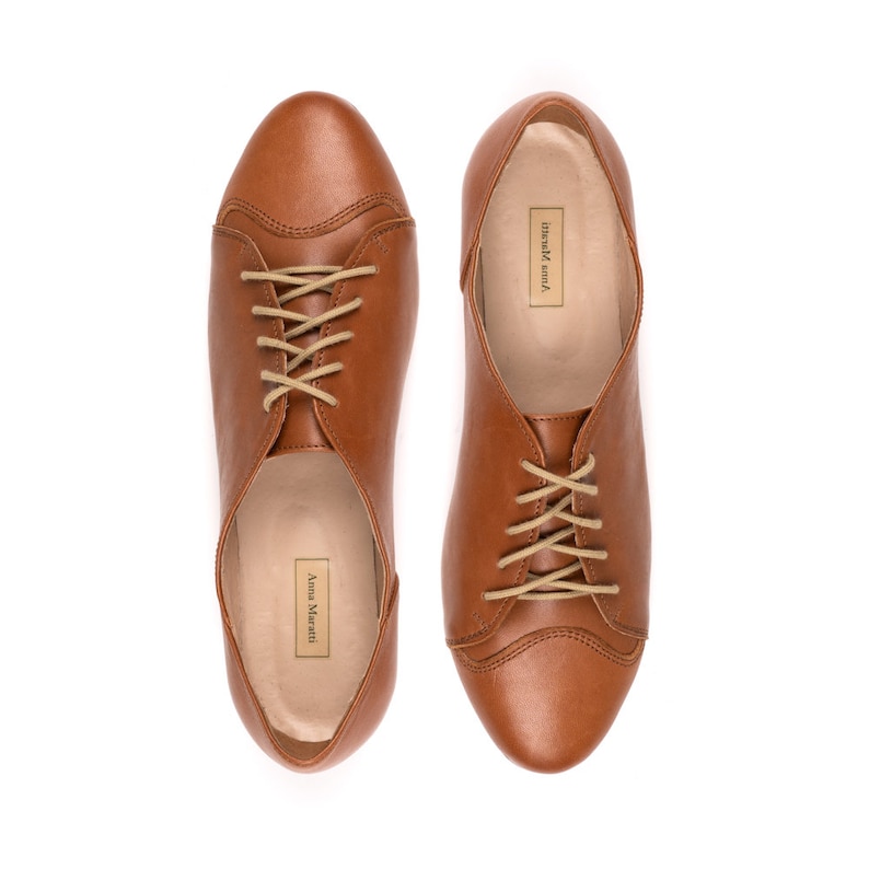 May include: A pair of brown leather lace-up shoes with tan laces. The shoes have a rounded toe and a low heel. The brand name "Anna Mariotti" is printed on the inside of the shoe.