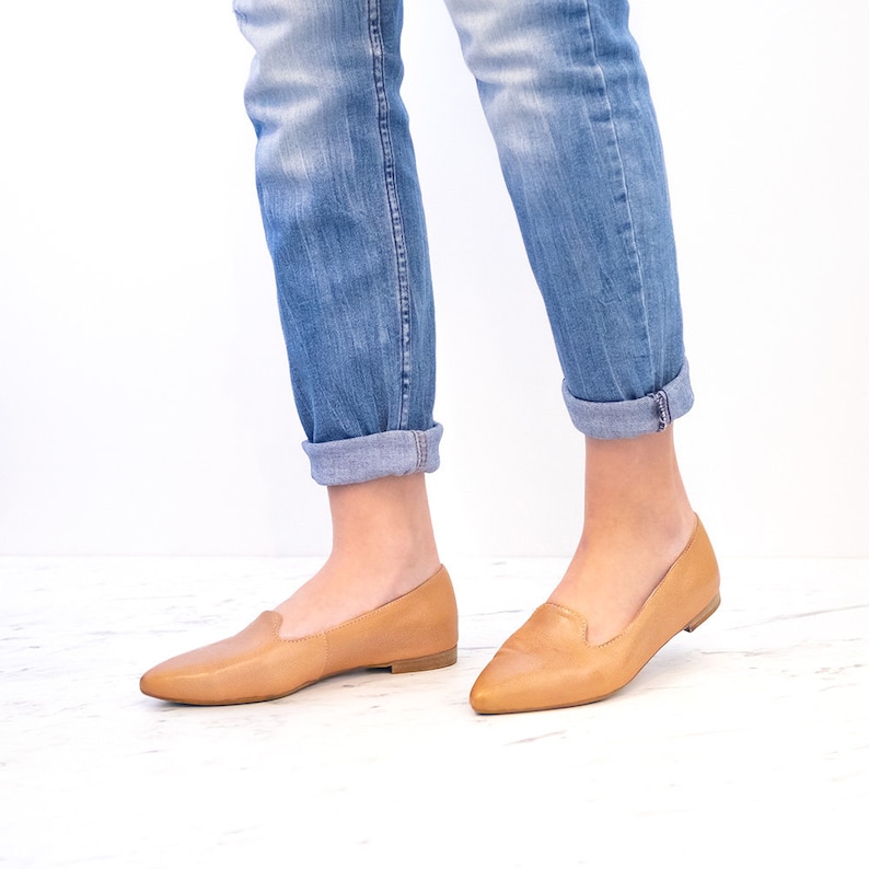 Op de afbeelding: Een paar beige leren spitse ballerina's met een lage hak. De schoenen worden gedragen door iemand die een blauwe jeans draagt.