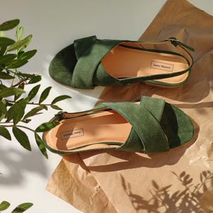 Pode incluir: Um par de sand&aacute;lias de camur&ccedil;a verde com tiras cruzadas e tiras no tornozelo. As sand&aacute;lias s&atilde;o exibidas em uma superf&iacute;cie marrom clara, com um ramo de folhas verdes ao fundo. A marca "Anna Marani" &eacute; vis&iacute;vel.