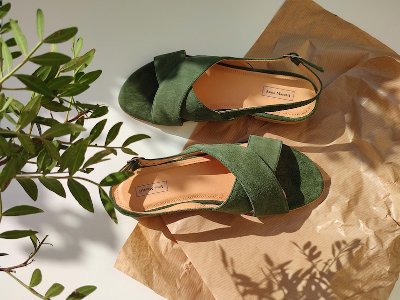 Pode incluir: Um par de sand&aacute;lias de camur&ccedil;a verde com tiras cruzadas e tiras no tornozelo. As sand&aacute;lias s&atilde;o exibidas em uma superf&iacute;cie marrom clara, com um ramo de folhas verdes ao fundo. A marca "Anna Marani" &eacute; vis&iacute;vel.