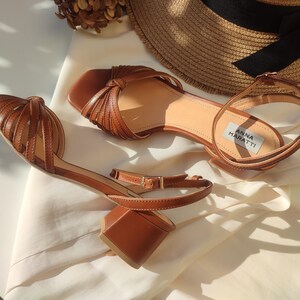 Puede incluir: Sandalias de cuero marr&oacute;n con tac&oacute;n de bloque y dise&ntilde;o de tiras. Las sandalias tienen correas en el tobillo con cierre de hebilla. El nombre de la marca "ANNA MARATTI" es visible en la plantilla. Un sombrero de paja est&aacute; en el fondo.