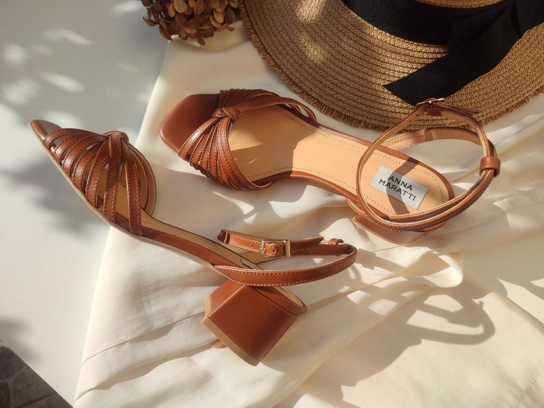 Puede incluir: Sandalias de cuero marr&oacute;n con tac&oacute;n de bloque y dise&ntilde;o de tiras. Las sandalias tienen correas en el tobillo con cierre de hebilla. El nombre de la marca "ANNA MARATTI" es visible en la plantilla. Un sombrero de paja est&aacute; en el fondo.