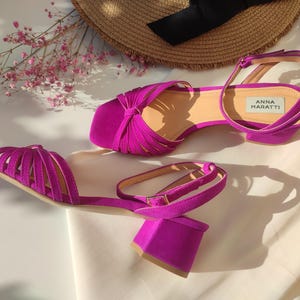 Może przedstawiać: Para żywych fuksjowych sandał&oacute;w z klockowym obcasem i paskiem na kostkę. Sandały mają pasek na stopie, a na wkładce widnieje nazwa marki "ANNA MARATTI". Słomkowy kapelusz i r&oacute;żowe kwiaty w tle.