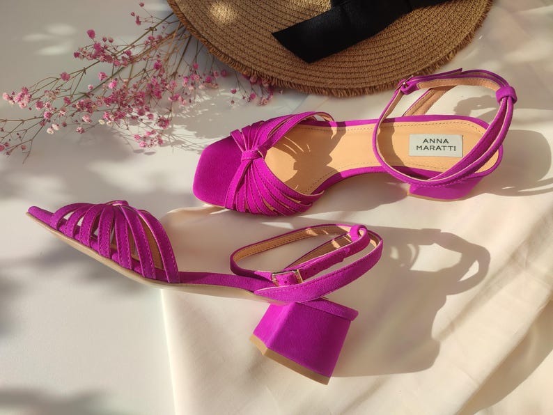 Może przedstawiać: Para żywych fuksjowych sandał&oacute;w z klockowym obcasem i paskiem na kostkę. Sandały mają pasek na stopie, a na wkładce widnieje nazwa marki "ANNA MARATTI". Słomkowy kapelusz i r&oacute;żowe kwiaty w tle.