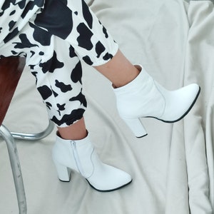 May include: White leather ankle boots with a block heel. The boots have a pointed toe and a zipper on the side. The boots are worn by a person wearing black and white cow print pants.