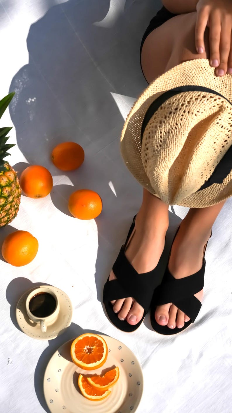 Pode incluir: Um chap&eacute;u de palha, sand&aacute;lias pretas e laranjas est&atilde;o dispostos sobre uma superf&iacute;cie branca. Uma x&iacute;cara de caf&eacute; e um prato com fatias de laranja tamb&eacute;m est&atilde;o presentes. Um abacaxi &eacute; vis&iacute;vel ao fundo, sugerindo um piquenique de ver&atilde;o.