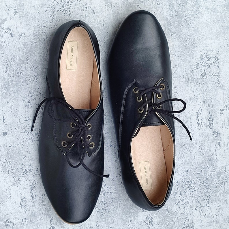 K&ouml;nnte beinhalten: Ein Paar schwarze Lederschn&uuml;rschuhe mit goldenen &Ouml;sen. Die Schuhe haben eine spitze Zehenpartie und einen niedrigen Absatz. Der Markenname "Anna Marani" ist auf der Innenseite der Schuhe sichtbar.